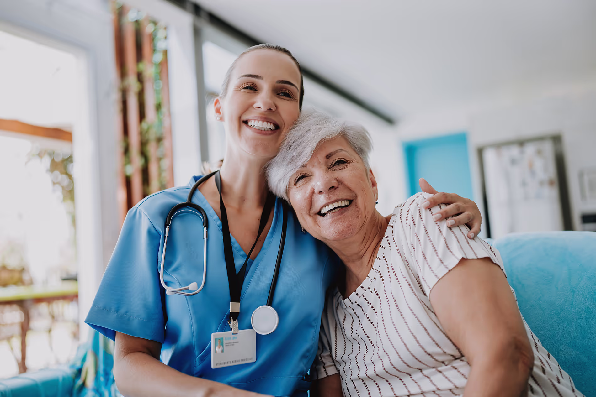 A nurse in blue scrubs smiles and hugs an elderly woman sitting beside her.