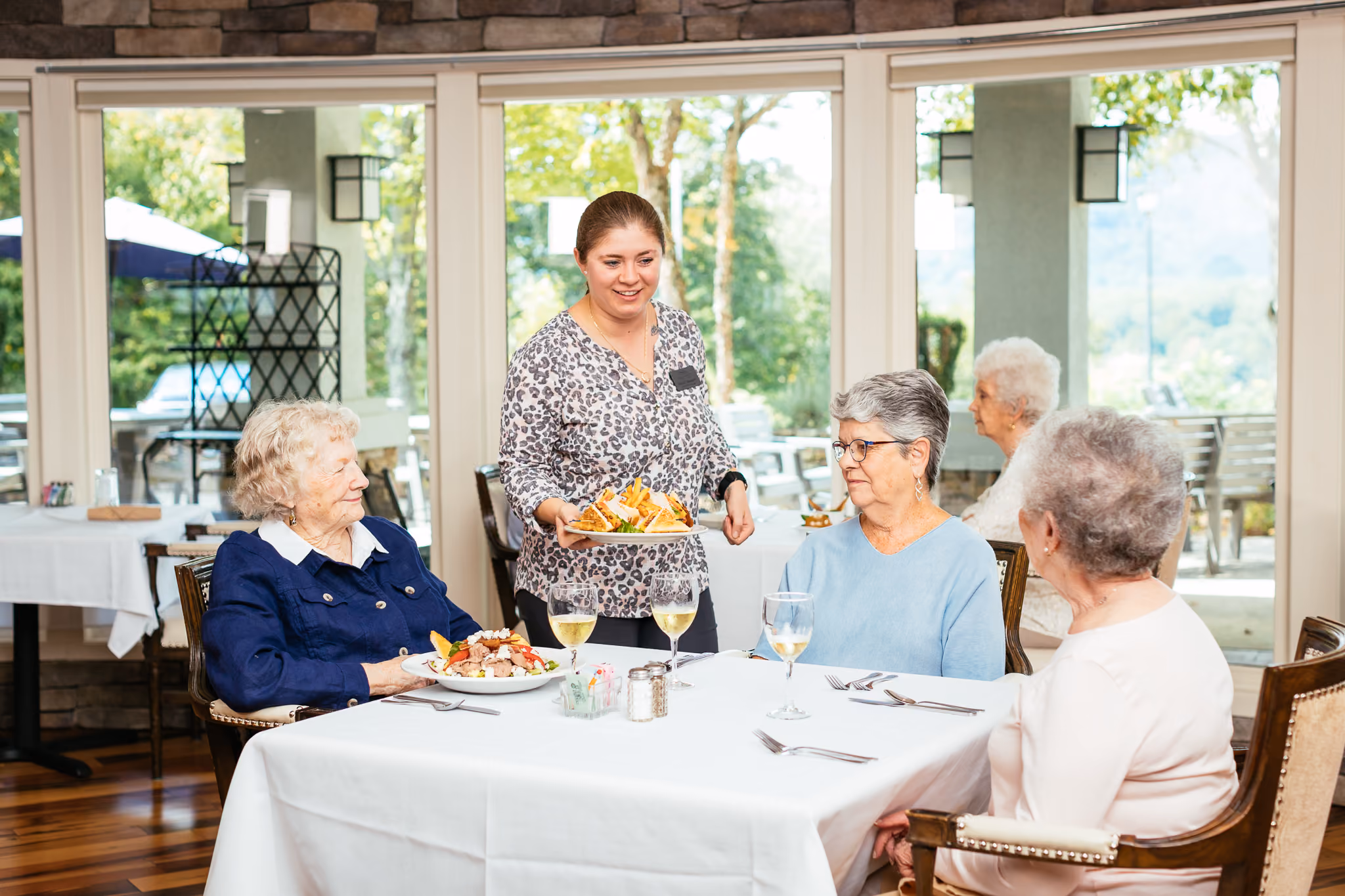 Server placing a plate of sandwich and fries before three elderly women dining at a white-clothed table.