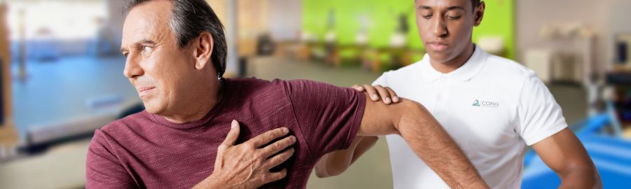 A physical therapist assists a male patient with shoulder discomfort in a clinic setting. The therapist is gently supporting the patient's arm while he expresses concern.