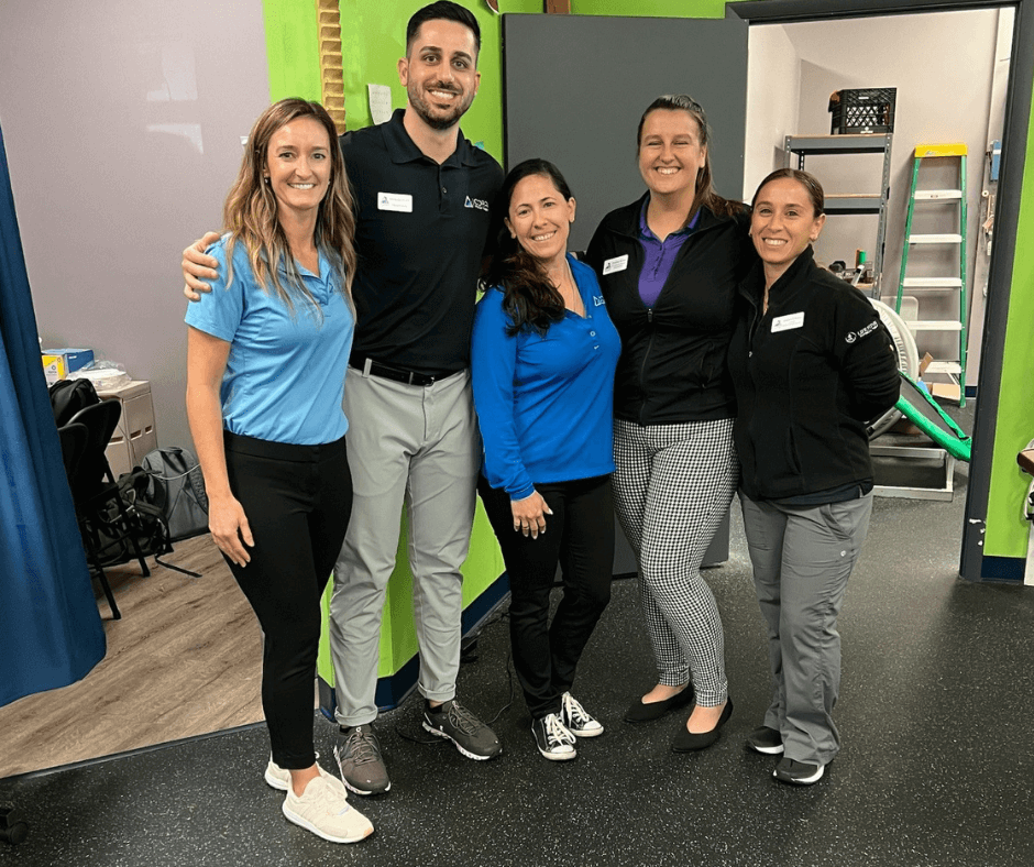 A group of five professionals posing together in an office environment. They are standing in front of a green wall, dressed in business casual attire, and smiling at the camera. Tools and equipment are visible in the background.