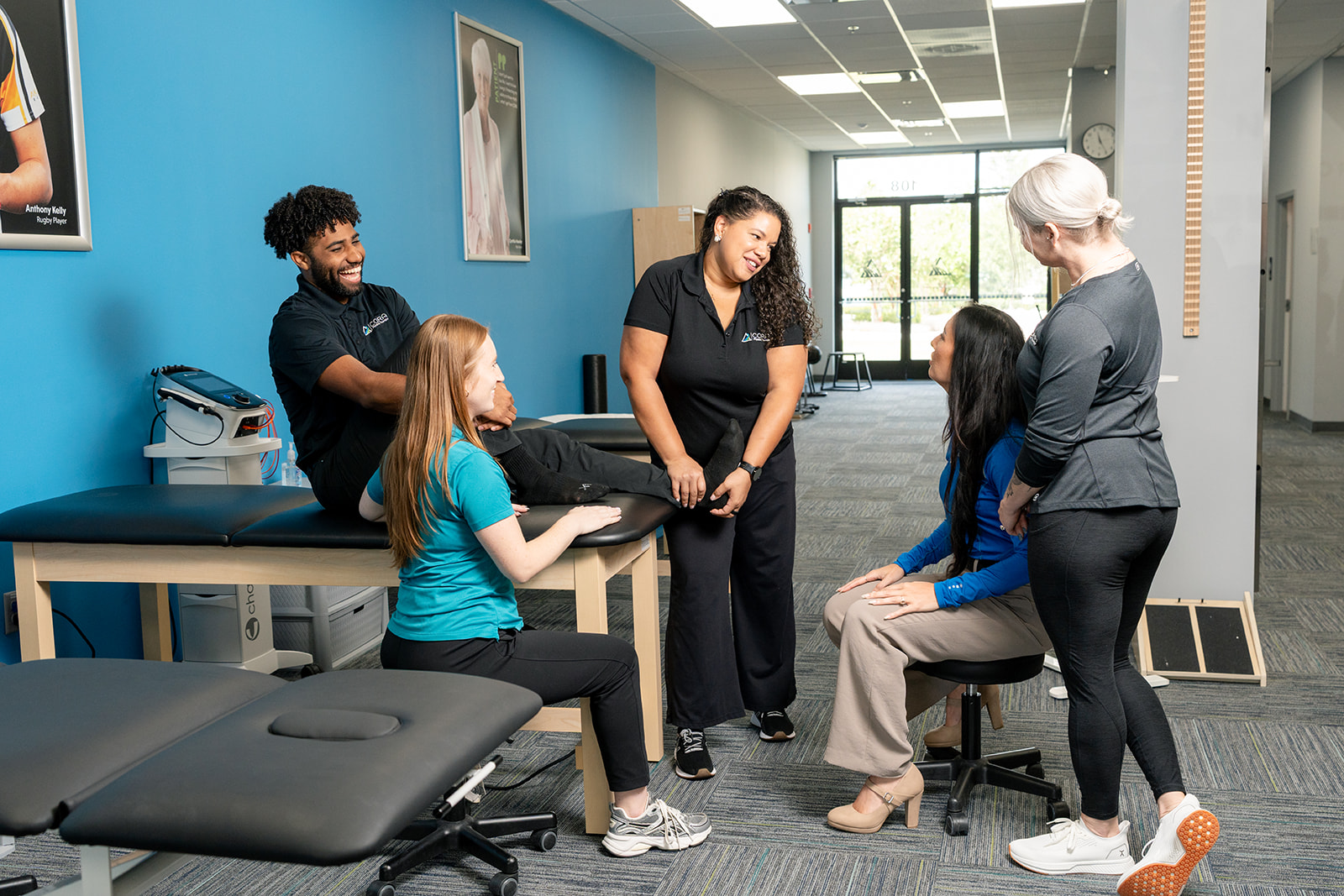A physical therapist assessing a patient's leg while she lies on a treatment table in a bright, modern clinic. The patient is wearing a blue shirt and black leggings.