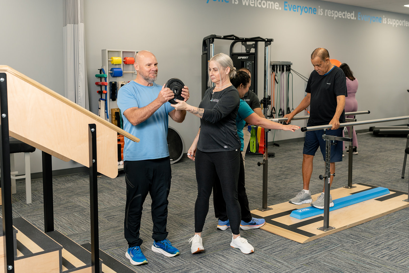 A woman assisting an older man with strength training using a small green dumbbell in a rehabilitation setting. The man is smiling and extending his arm while sitting on a treatment table.