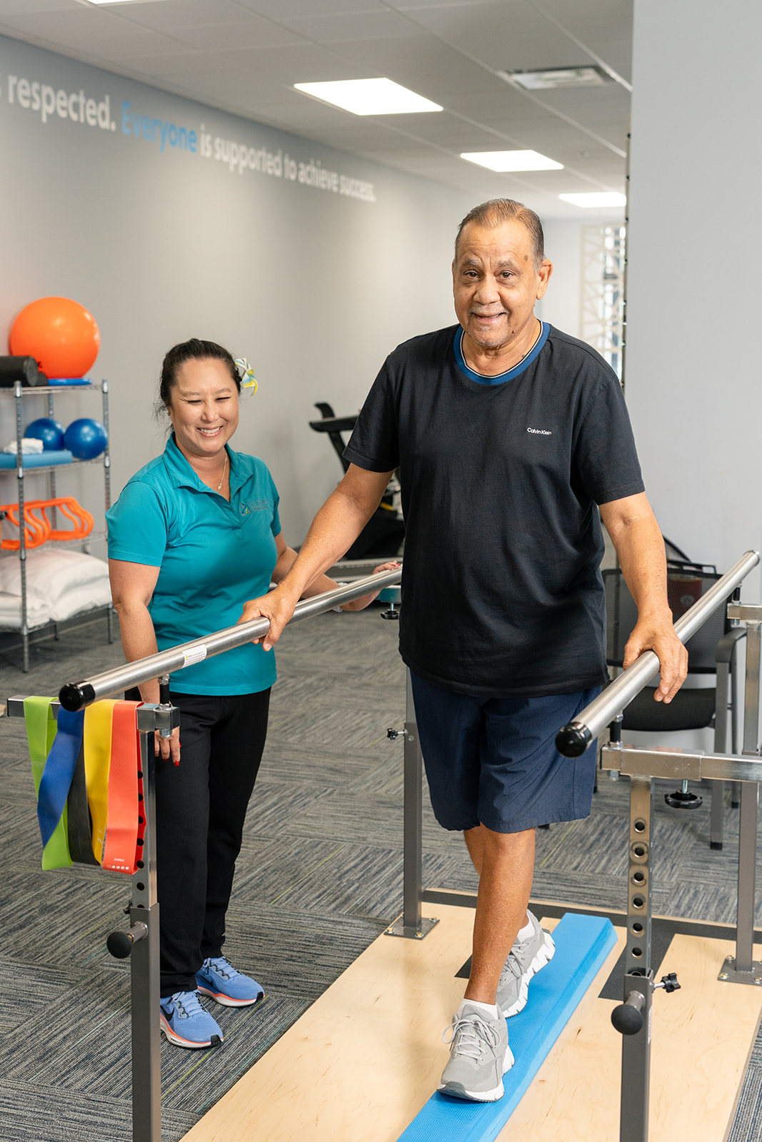 A female physical therapist standing with a tablet, talking to an elderly male patient seated on an examination table, in a bright therapy room with plants and medical supplies.