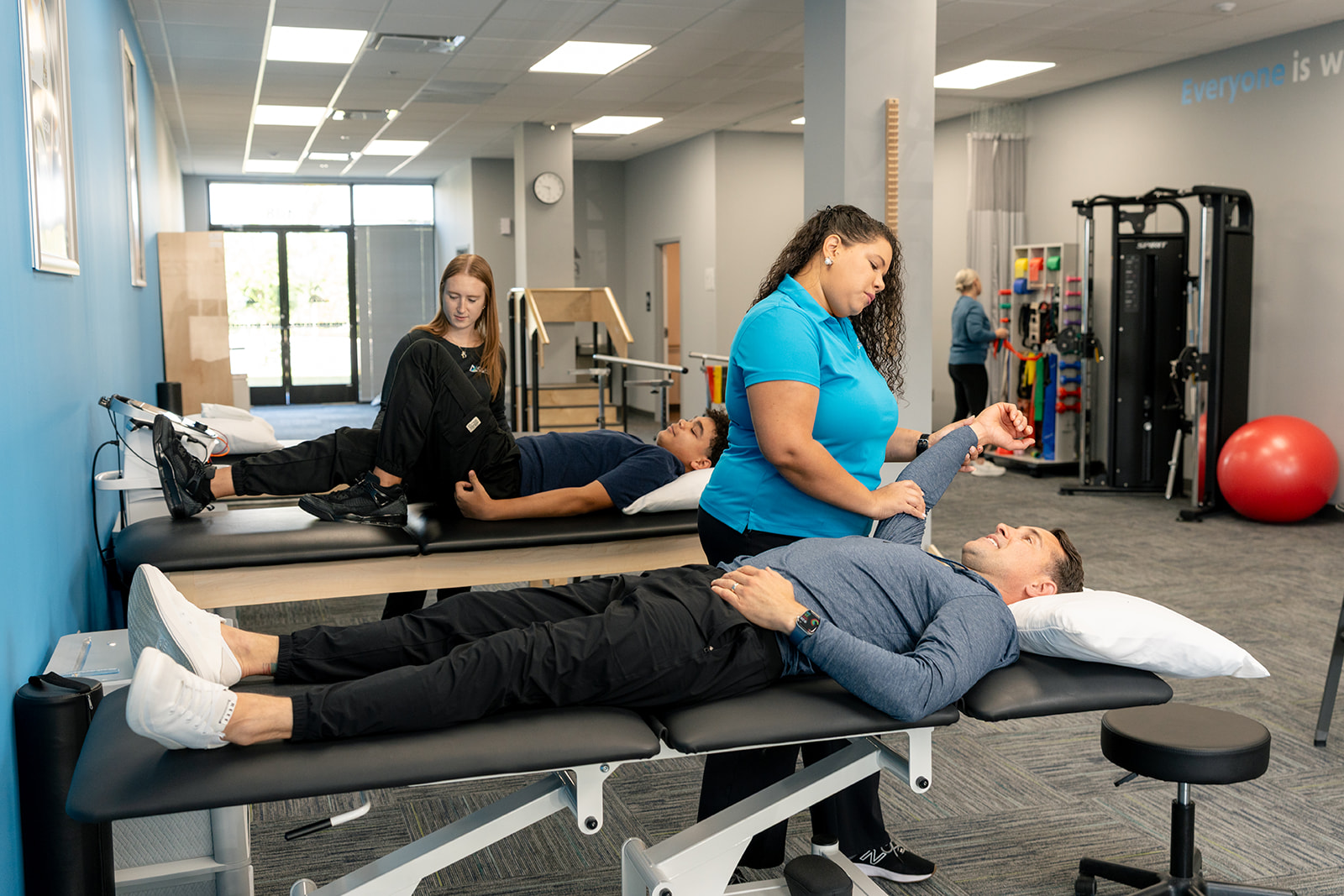 A physical therapist assists a male patient with shoulder discomfort in a clinic setting. The therapist is gently supporting the patient's arm while he expresses concern.