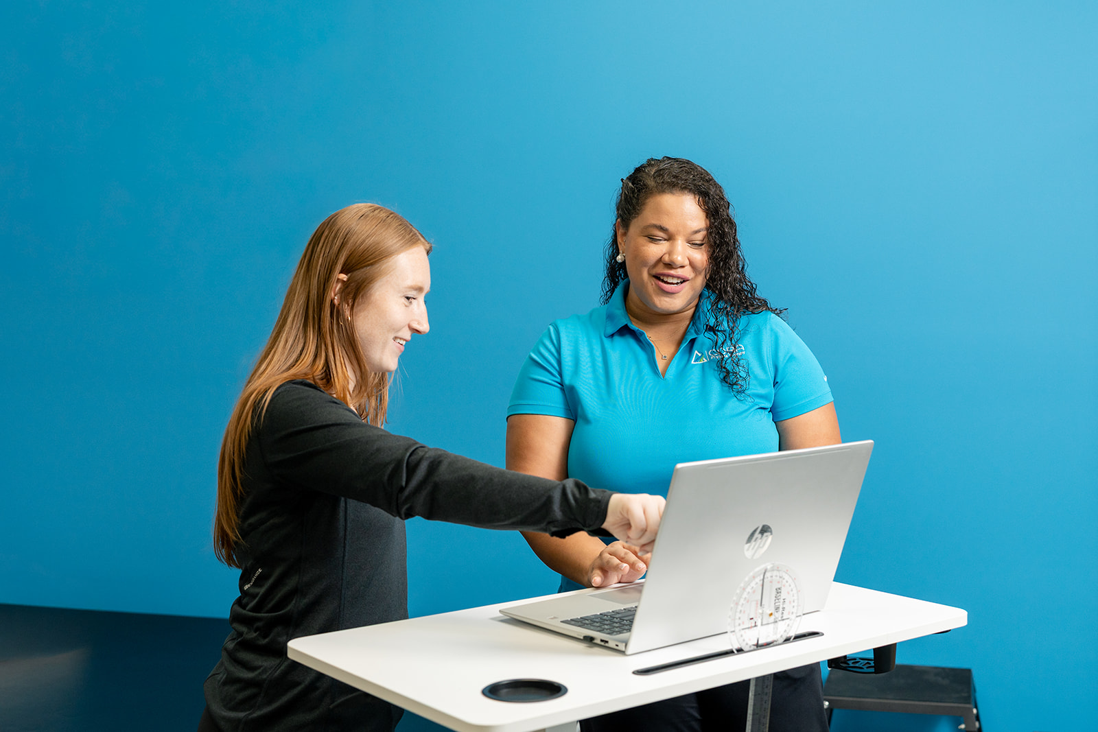 A woman and a young girl interact at a table, with the woman pointing to a chalkboard while the girl holds a marker. Colorful art supplies are visible on the table in a bright, modern learning space.