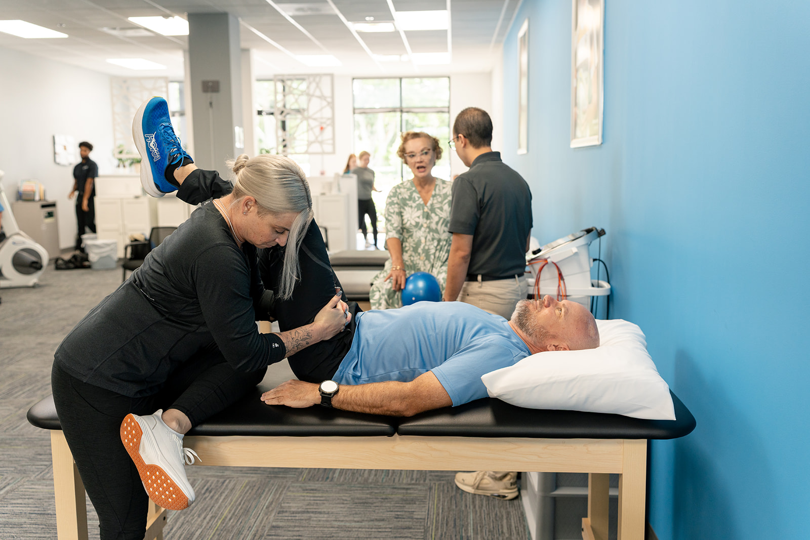 A group of people sitting on the floor in a training session, with one person demonstrating a technique on a lying participant. Others are taking notes and referring to handouts.