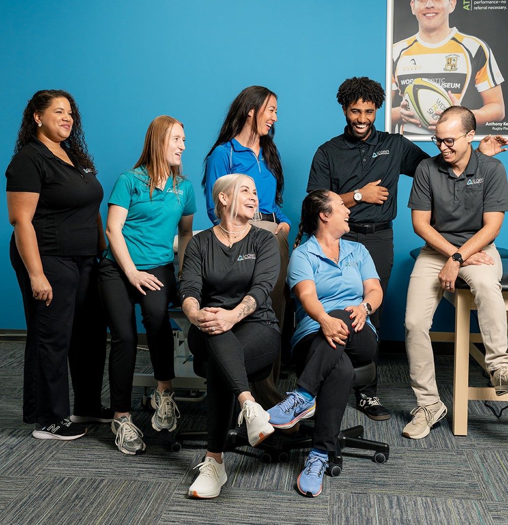 A group of five professionals posing together in an office environment. They are standing in front of a green wall, dressed in business casual attire, and smiling at the camera. Tools and equipment are visible in the background.