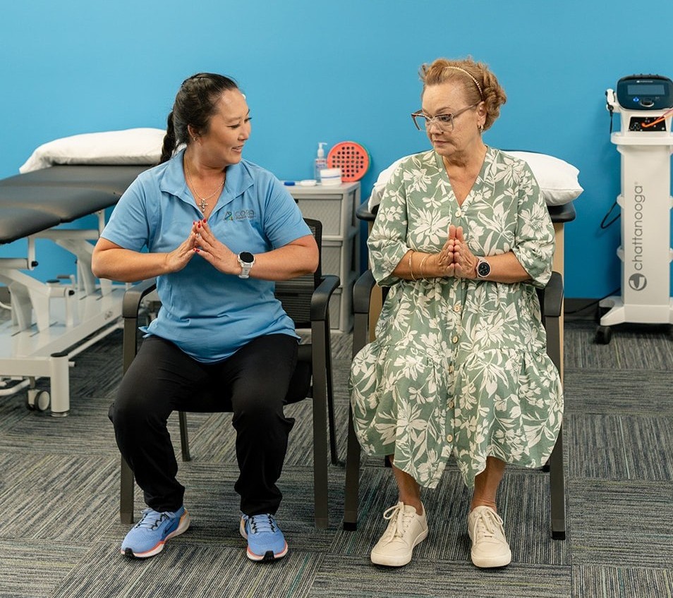 A woman and a girl sitting on the floor, both mimicking facial expressions with their hands on their faces. The setting is a playful room with exercise balls and children's bikes in the background.