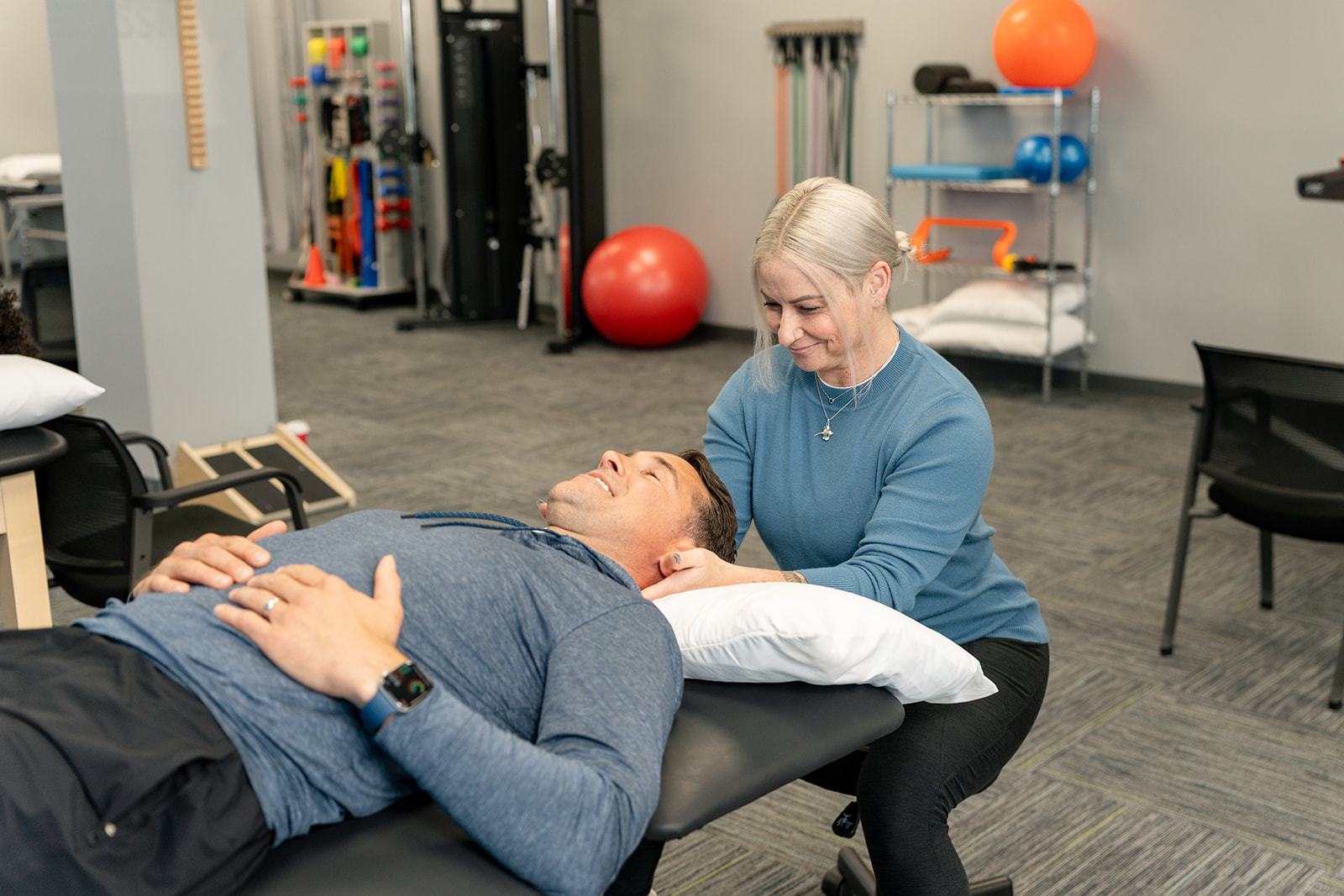 A physical therapist assists a male patient with shoulder discomfort in a clinic setting. The therapist is gently supporting the patient's arm while he expresses concern.