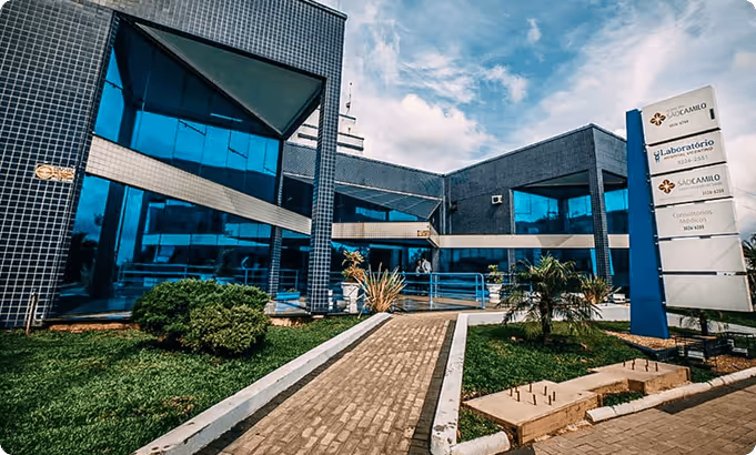 Vista da fachada de um prédio administrativo com paredes de azulejos escuros e janelas espelhadas refletindo o céu azul, jardim com arbustos e caminho de blocos de concreto levando à entrada.