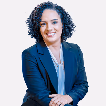 Mulher sorridente com cabelo cacheado veste blazer azul-marinho e blusa branca, sentada com braços cruzados.