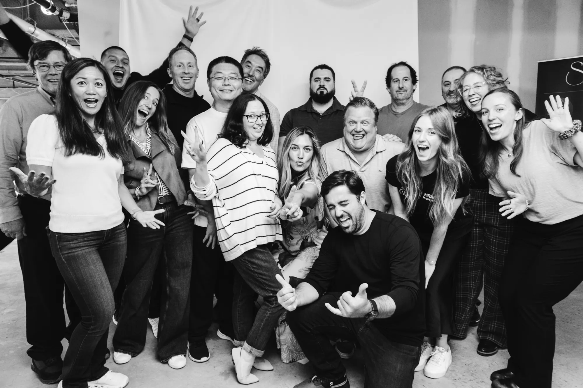 Group of happy people posing energetically indoors, some making peace signs and playful gestures.