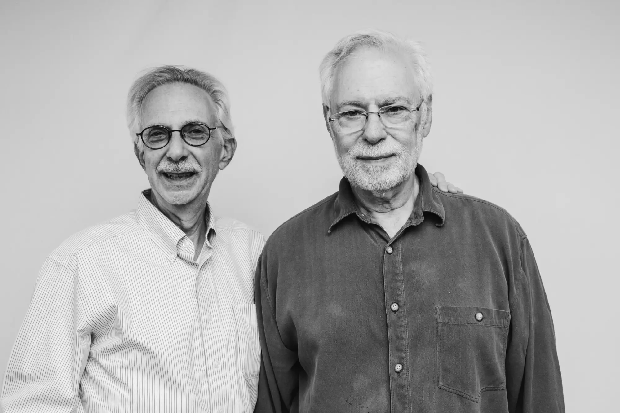 Two older men standing side by side, smiling, one with glasses and a striped shirt, the other with glasses and a button-up shirt.
