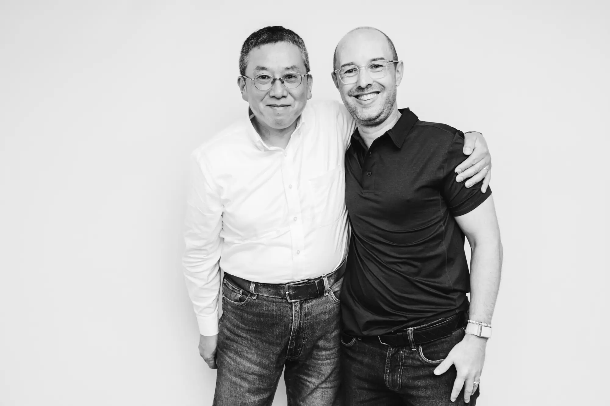 Two men standing closely with arms around each other smiling against a plain background.