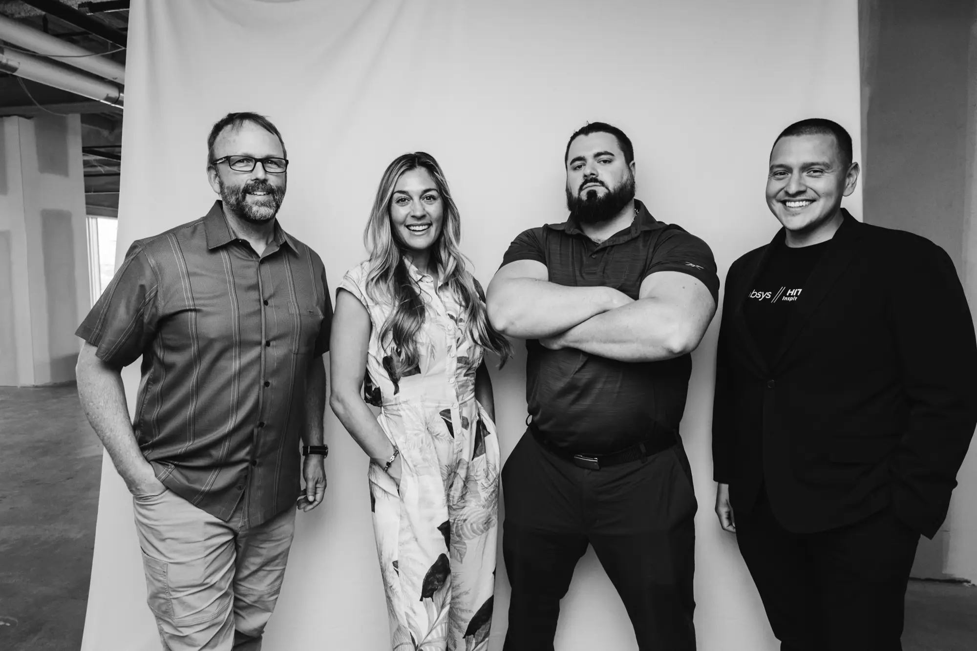 Black and white photo of four adults standing together smiling, two men and a woman with relaxed poses, one man crossing his arms.