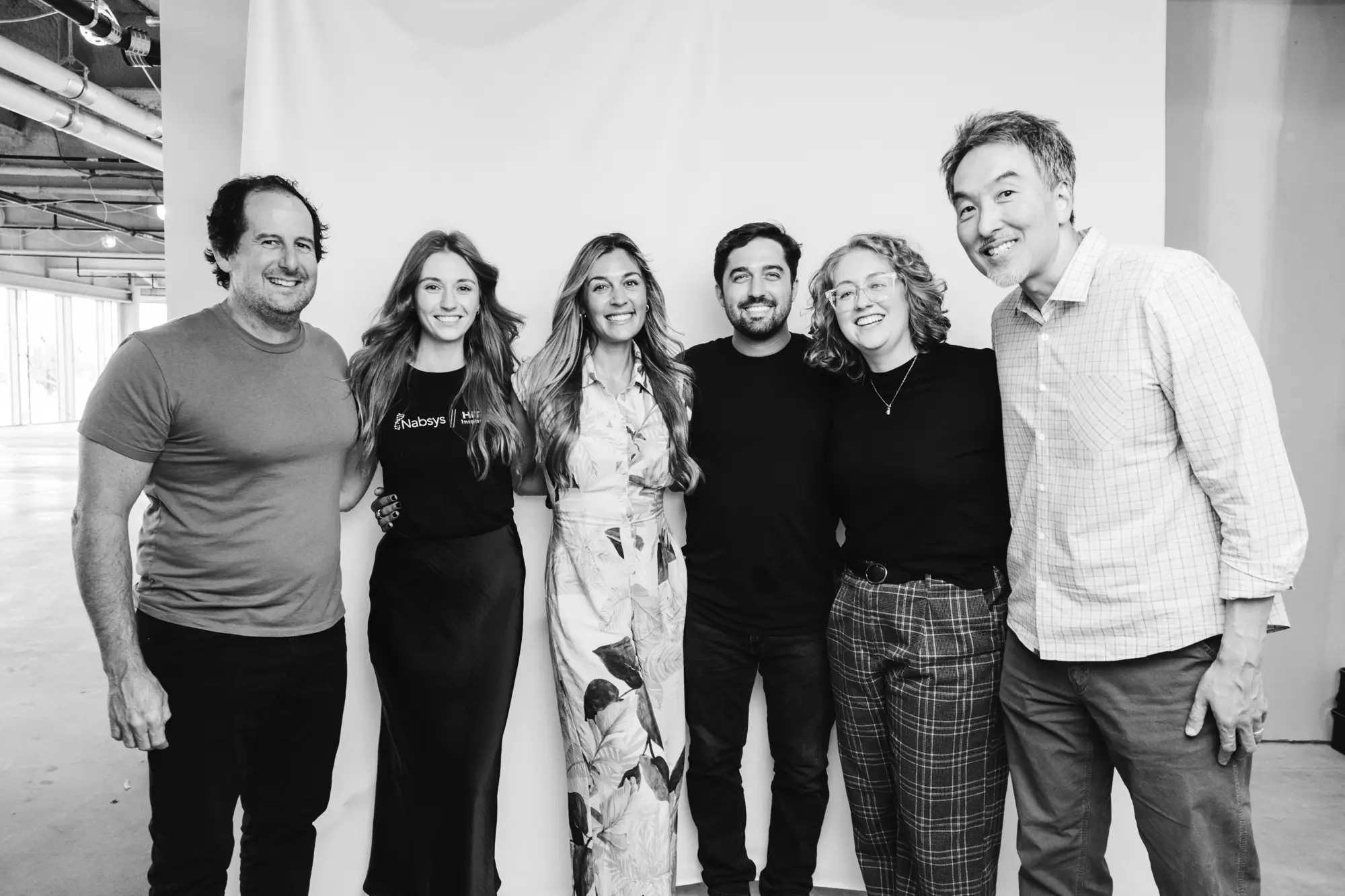 Six diverse people standing closely together smiling in a casual indoor setting with a plain backdrop.