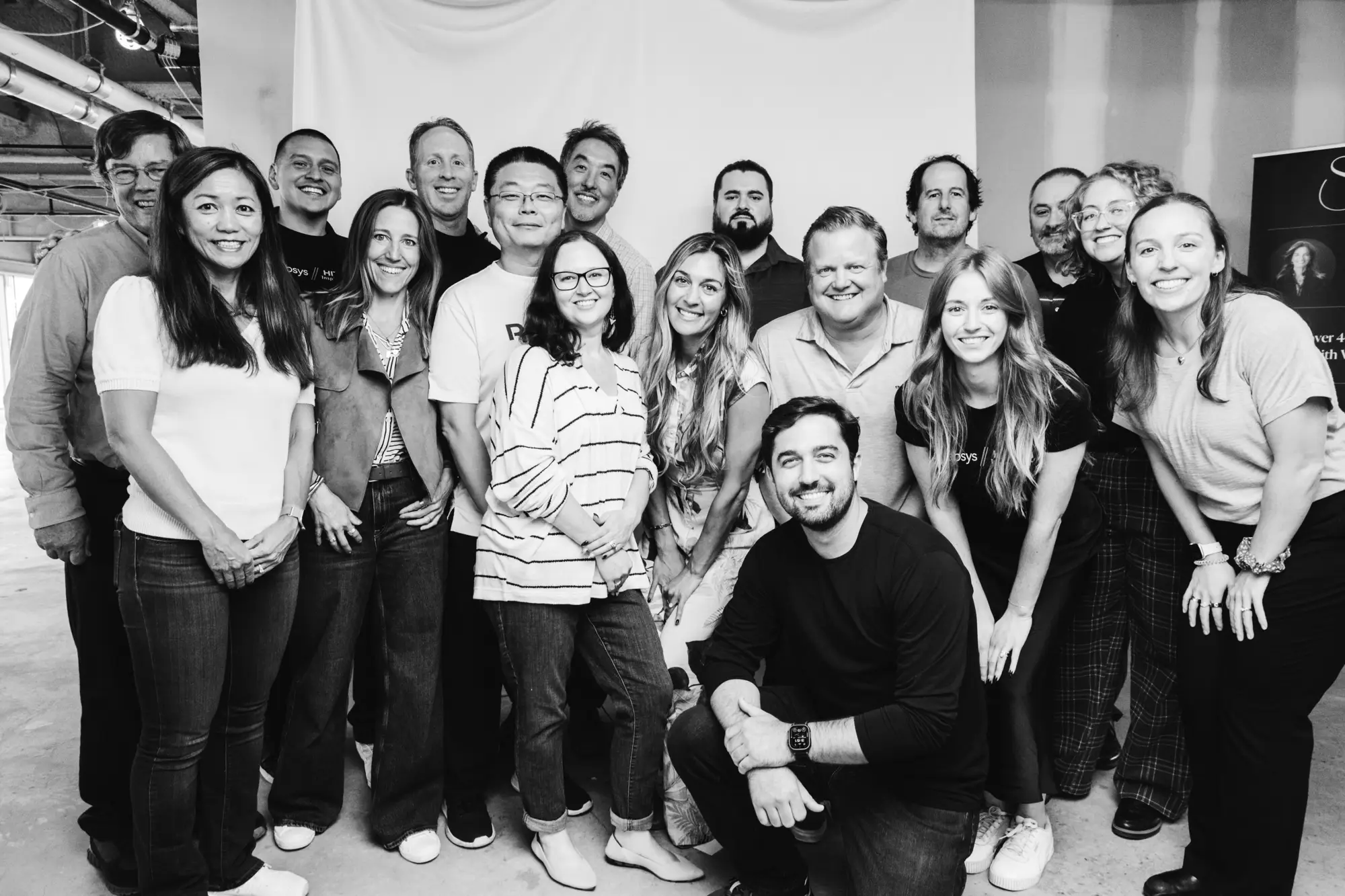 Black and white group photo of 16 smiling people posing indoors in a casual setting.
