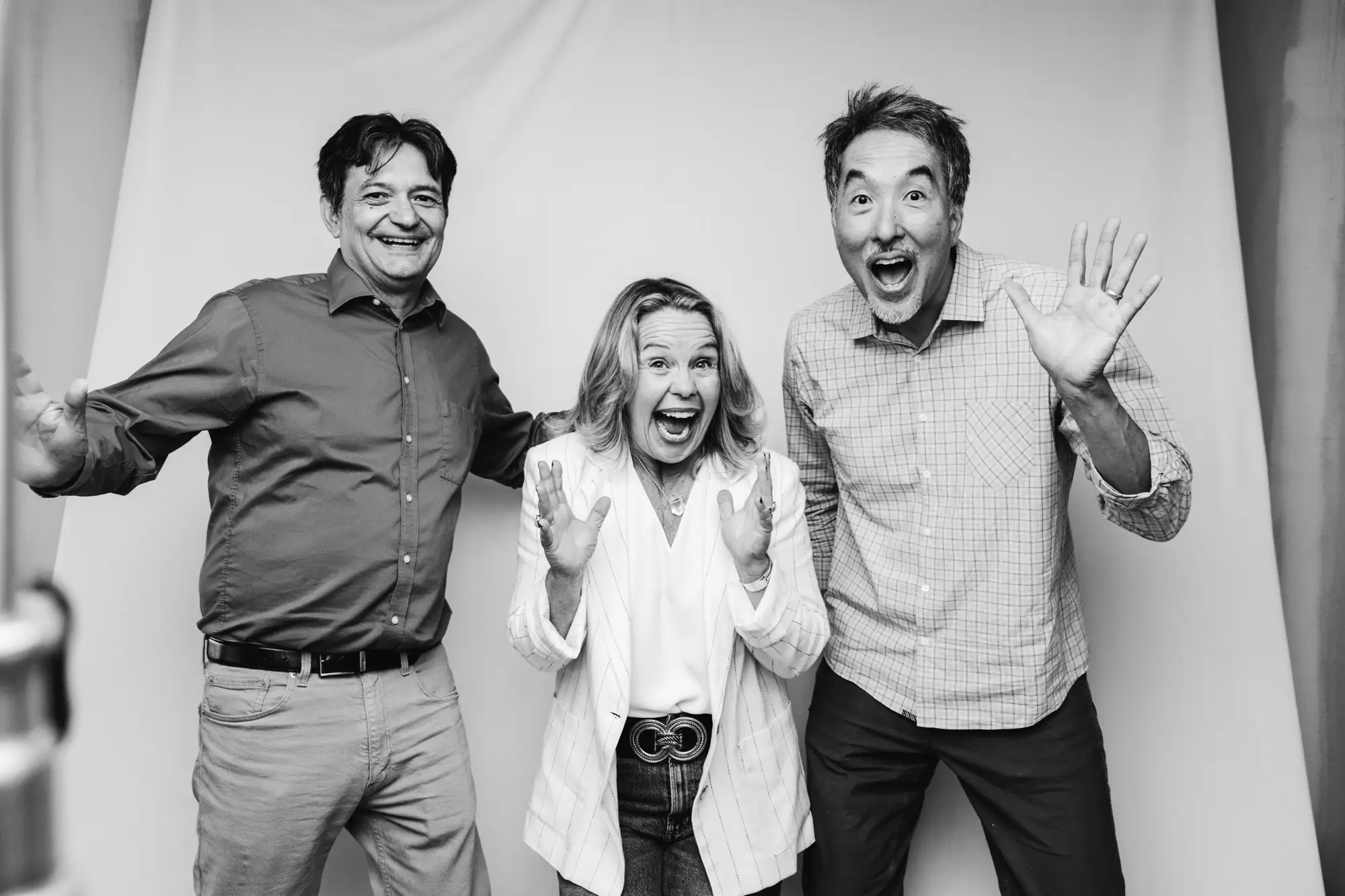 Three adults smiling and posing excitedly in front of a plain backdrop, two men and one woman in the center.