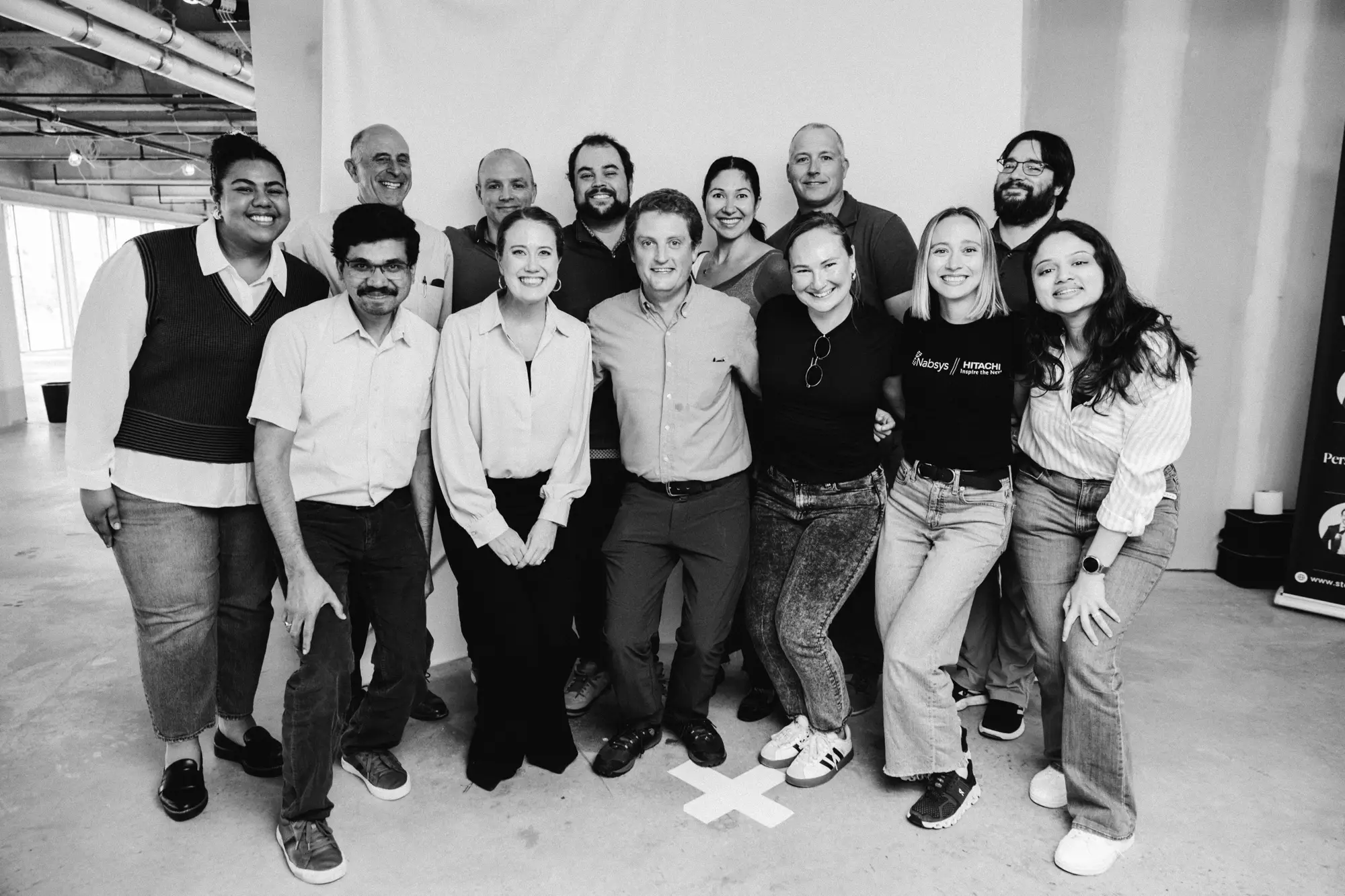 Group of thirteen diverse colleagues standing indoors smiling together in a casual office space.