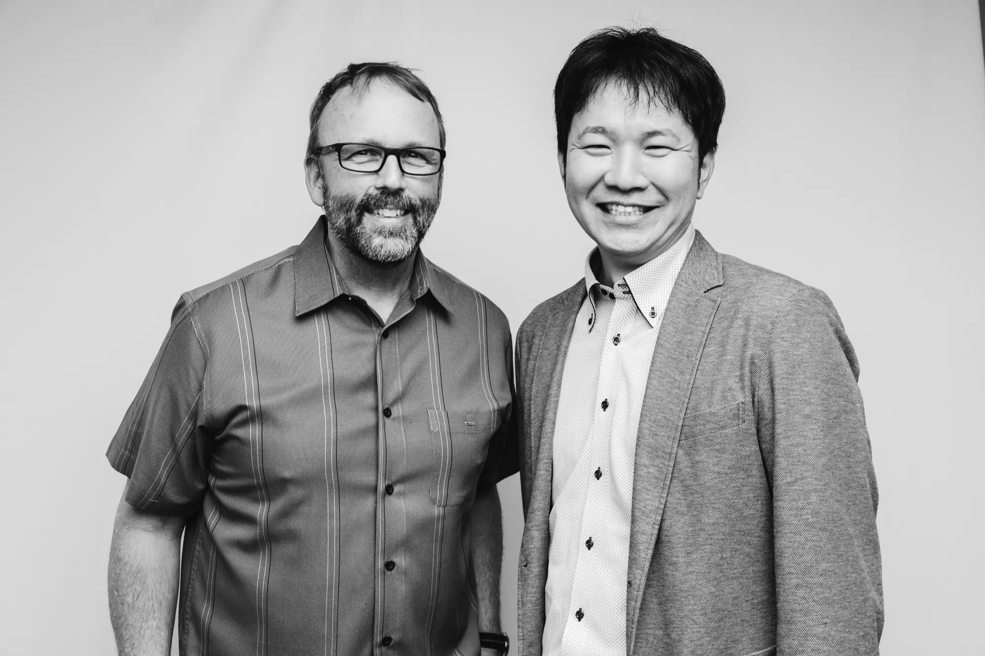 Two men smiling; the man on the left wears glasses and a striped short-sleeve shirt, and the man on the right wears a blazer over a buttoned shirt.