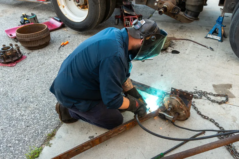 Technician performs welding and fabrication on metal frame near disassembled truck axle and brake components.