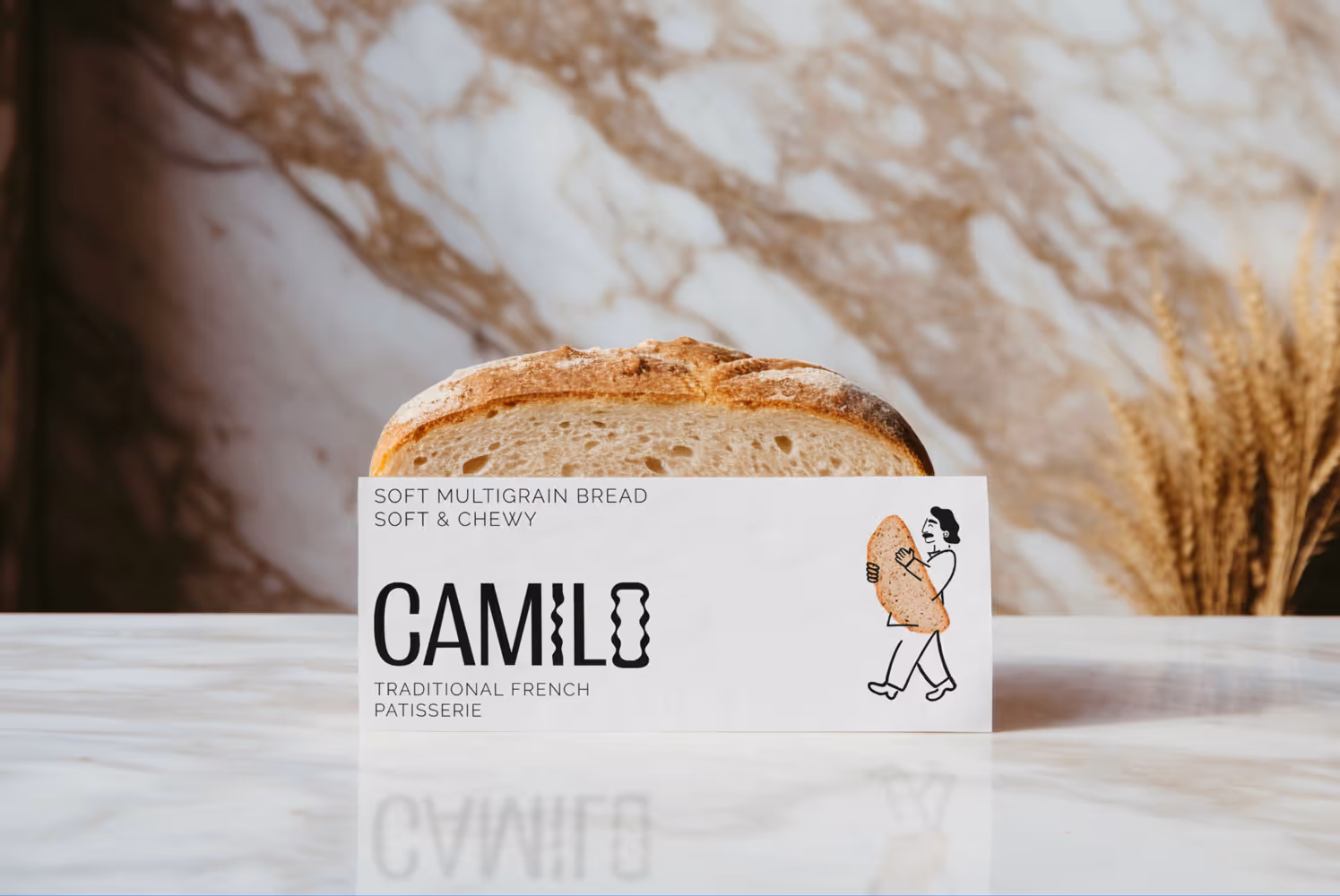 Hogaza de pan multigrano suave detrás de una etiqueta de la marca Camilo que dice "Pan multigrano suave, suave y tierno, pastelería francesa tradicional" con una figura de caricatura cargando una baguette