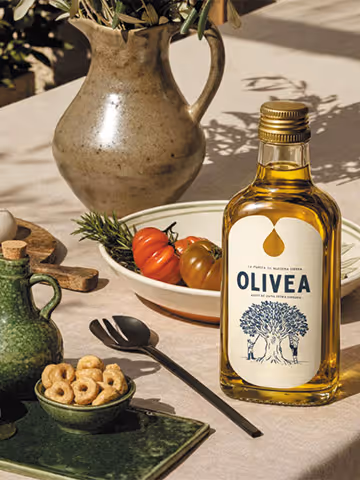 Botella de aceite de oliva Olivea sobre una mesa junto a un cuenco de tomates, un pequeño cuenco verde de aperitivos y jarras de cerámica.