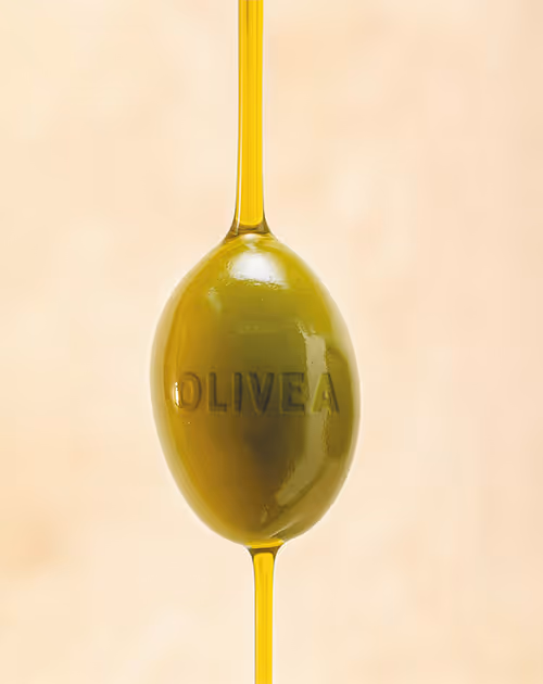 Primer plano de una aceituna verde con aceite de oliva goteando por arriba y por abajo sobre un fondo beige.