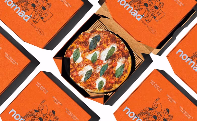 Vista superior de una pizza margarita con hojas de albahaca en una caja de pizza naranja y negra abierta, rodeada de cajas cerradas con la marca 'Nomad'