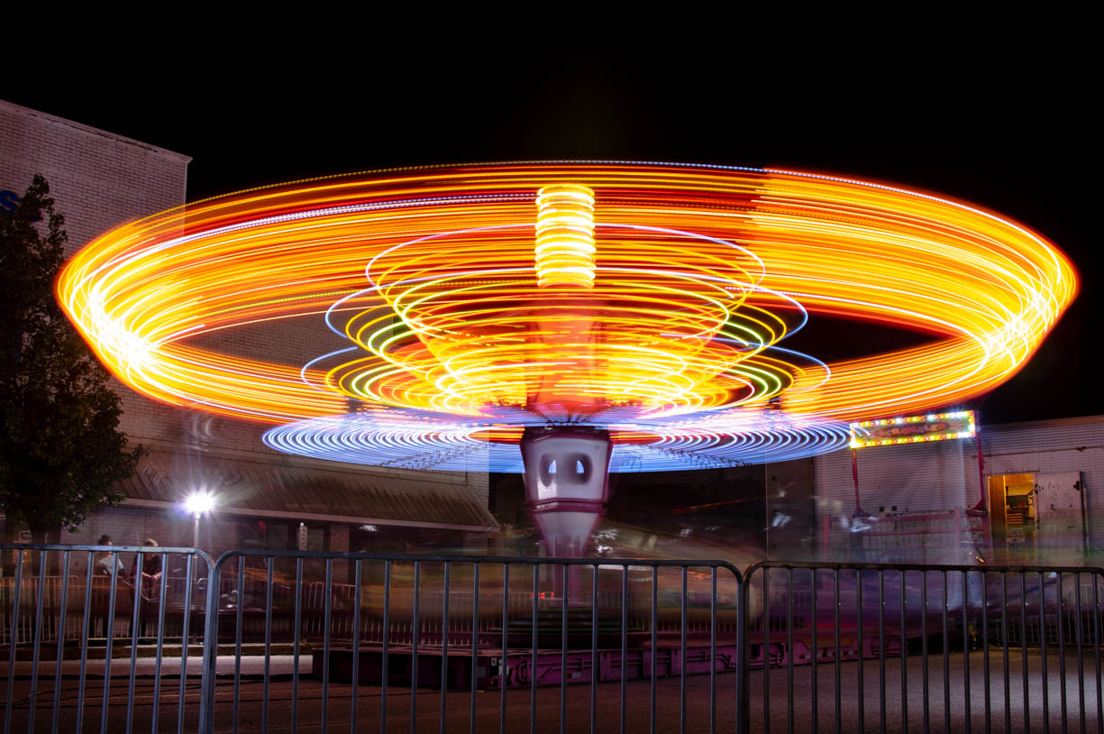 A long exposure shot of a spinning fair ride. The ride is orange, red, and blue