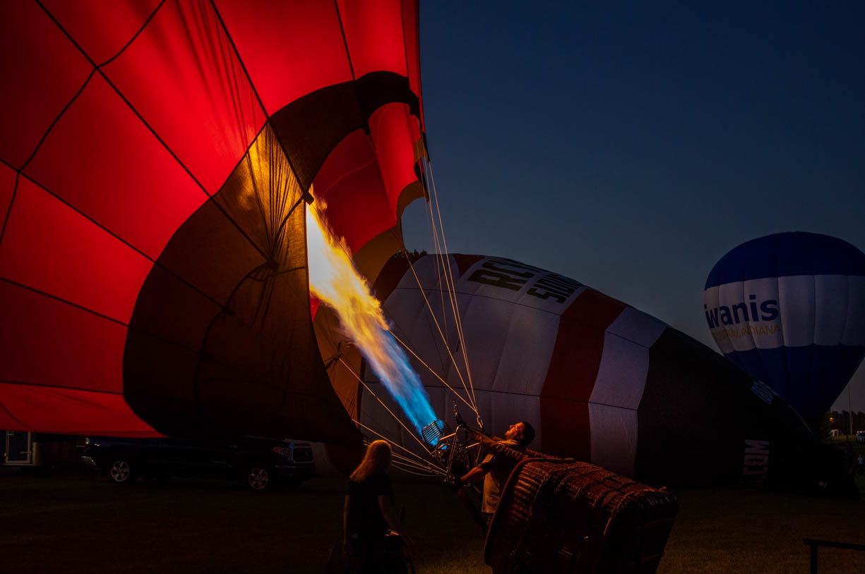 A hot air balloon being lit up during a hot air balloon night glow in central Indiana during summer 2022. The hot air balloon is bright red due to the blue and orange flame shooting up into the balloon. A man is holding the device that allows the balloon to gain lift. 