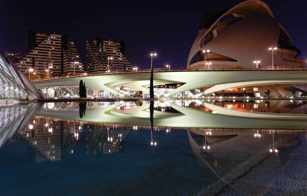 Some spectacular reflections of the modern architecture in the City of Arts and Sciences in Valencia, Spain. The reflections include the Imax theatre, modern apartment buildings and a modern bridge that is illuminated in green lights. The sky is purple and the water is blue.