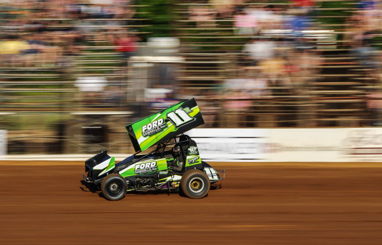 A green and black 410 sprint car racing in front of the grandstands at high speed. The background of the image is blurred but the car is in focus creating the sense of speed.