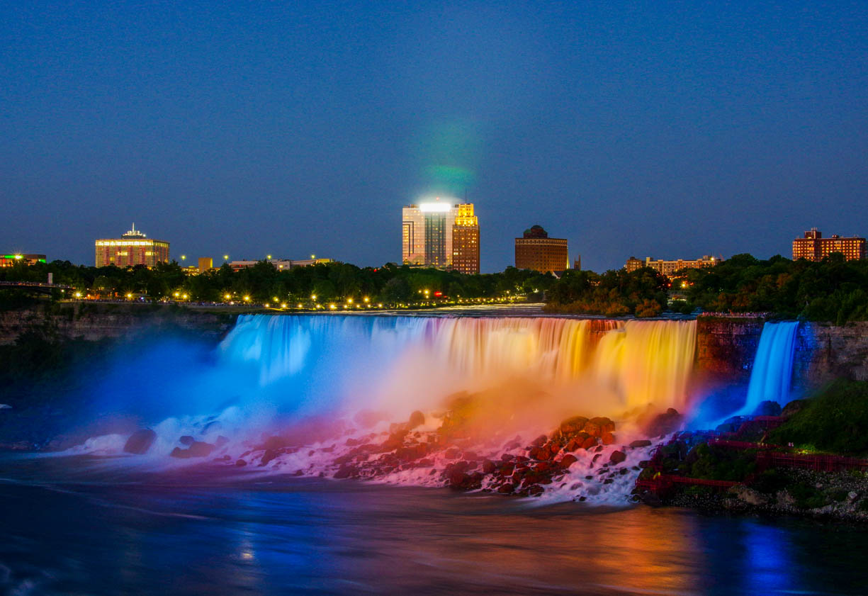 This image shows the American side of Niagara Falls at night. This image was captured from the Canadian side in Niagara Falls, Canada. This scenic location is a popular tourist attraction, especially at night because of the scenic light show that occurs every night at the falls. In this image of the falls, the waterfall is blue and orange because of lights that illuminate the falls from behind it.
