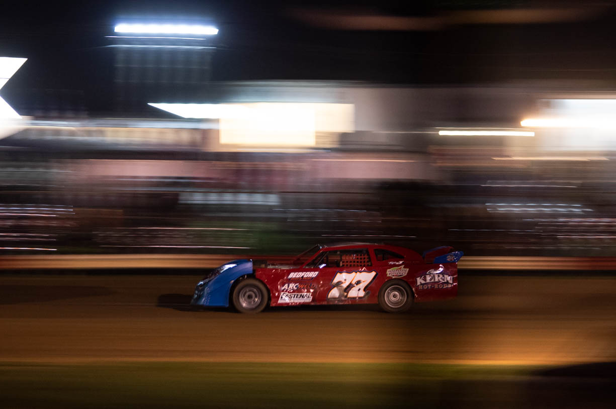 A red and blue stock car racing at high speeds at the Paragon Speedway in Bloomington, Indiana. The racecar is racing at high speeds in the night time, therefore, it is illuminated by the bright lights