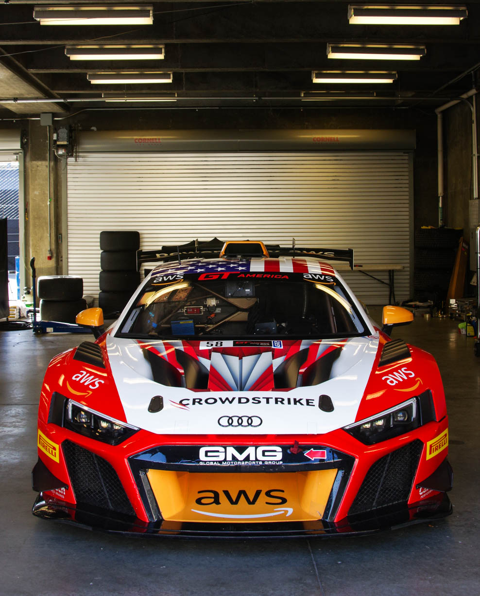 A race car Audi R8 photographed in its garage at the Indianapolis Motor Speedway during the 8 hour race in the fall of 2024. The car is bright orange and white with a beautiful American flag on the hood. and the top of the car.   