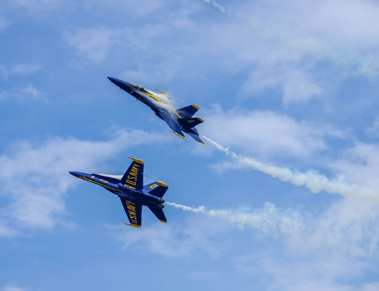 Two Blue Angels F-18 planes making an aerobatic maneuver in the sky during an airshow, the planes are extremely close together and are turning so quickly they are creating a band of moisture along the plane.