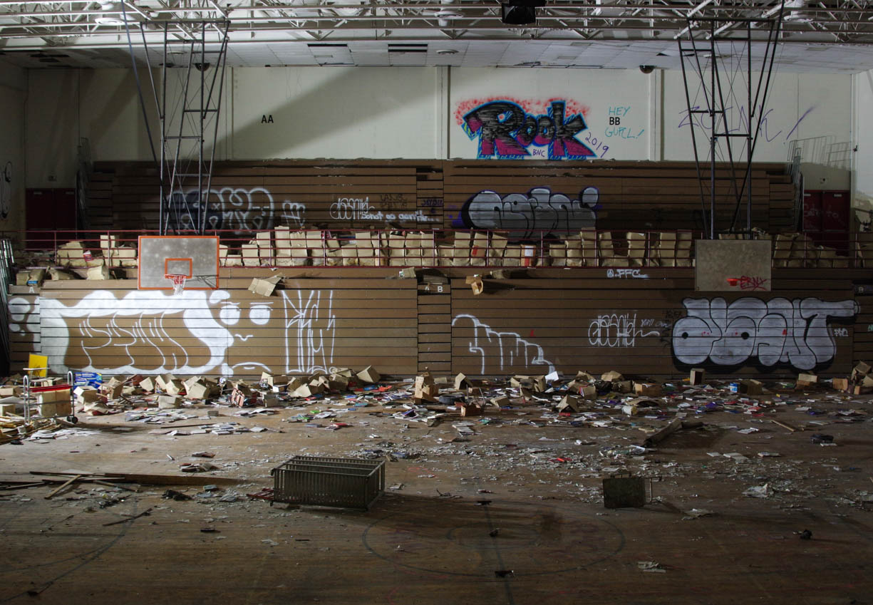 One of the gyms in the abandoned Horace Mann High School in northern Gary, Indiana. The photo shows two basketball hoops and bleachers in the background with boxed of books thrown everywhere all across the gym and some boxes are piled high in the background next to some graffiti at the top of the bleachers.