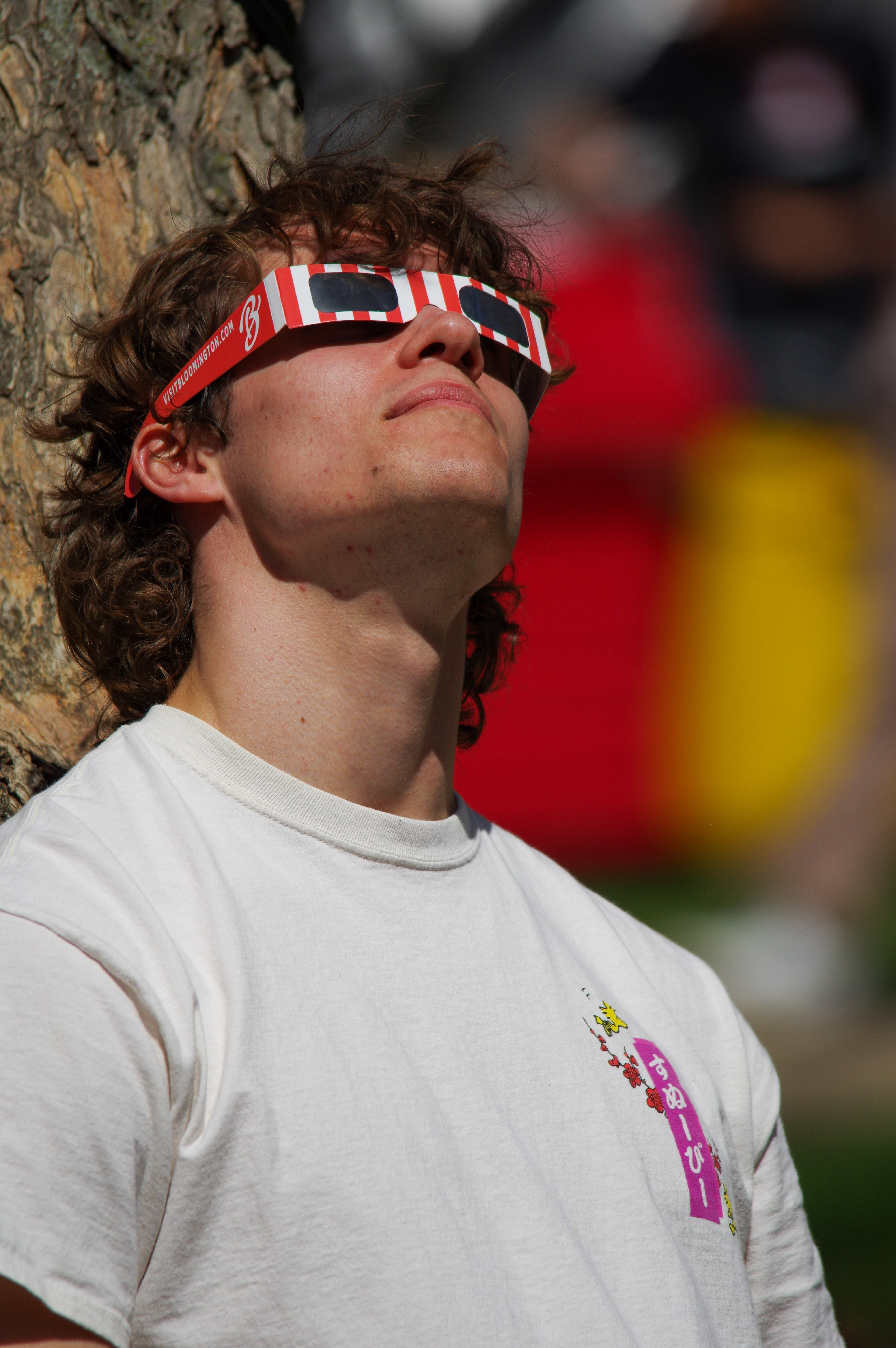 A young man looking up at the 2024 solar eclipse from Indiana University. The man is wearing red and white striped Visit Bloomington eclipse glasses and has brown hair. He is also good looking and is wearing a white shirt.
