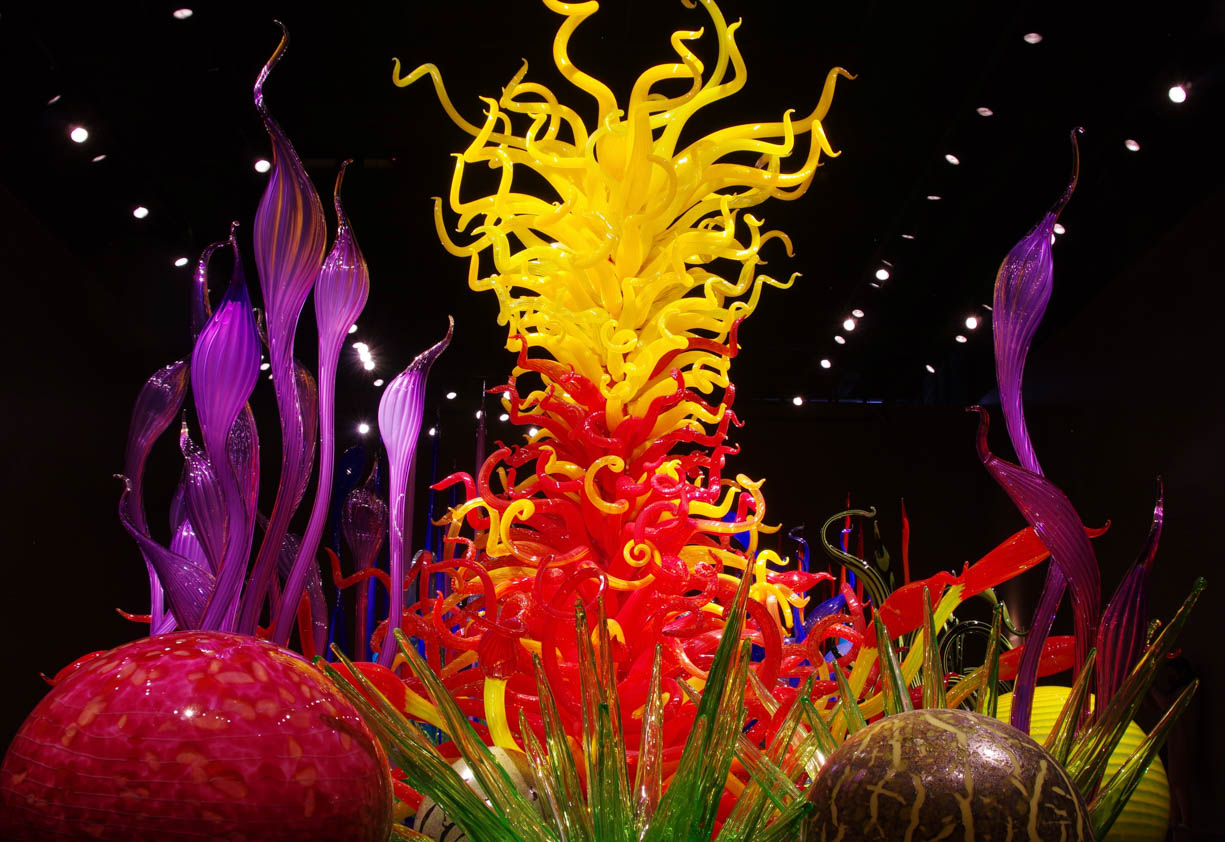 This image shows the popular glass centerpiece in the Chihuly Garden and Glass in Seattle, Washington. This scene can be viewed in person by standing in the first main room in Chihuly's Garden of Glass. This image was taken with a tripod to maximize viewing and resolution quality. This image was also taken during a west coast road trip in the summer of 2021. The glass structure looks like underwater coral in vibrant colors like red, yellow, green, and purple. The middle of the structure is red and yellow. 