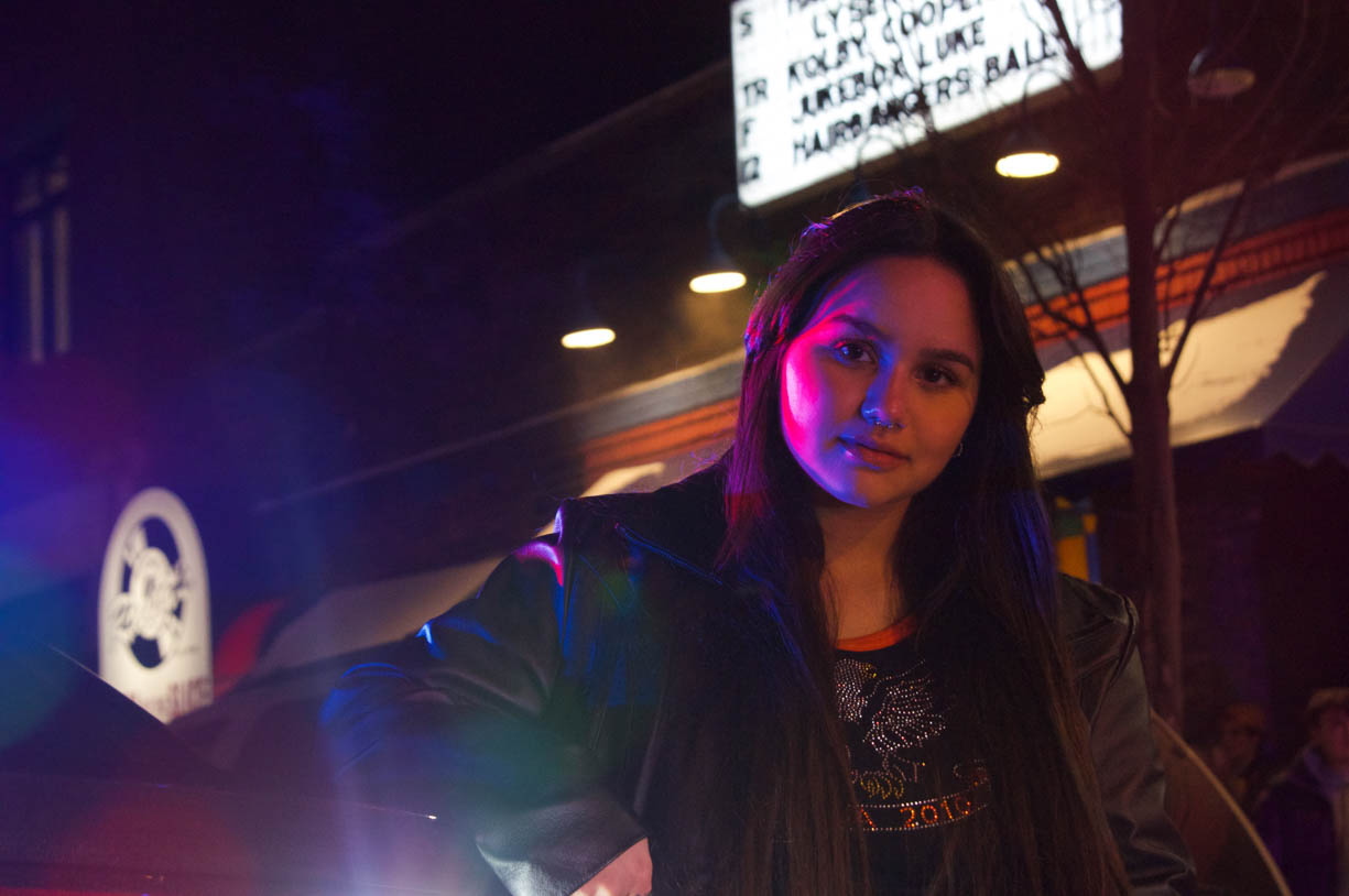 A young woman posing next to a police car as her face and body are being illuminated in red and blue from the colors atop the police car. She is wearing a bedazzled shirt and a black heather jacket and her hair is black and well as her brown eyes. The image only shows the upper half of her body. 