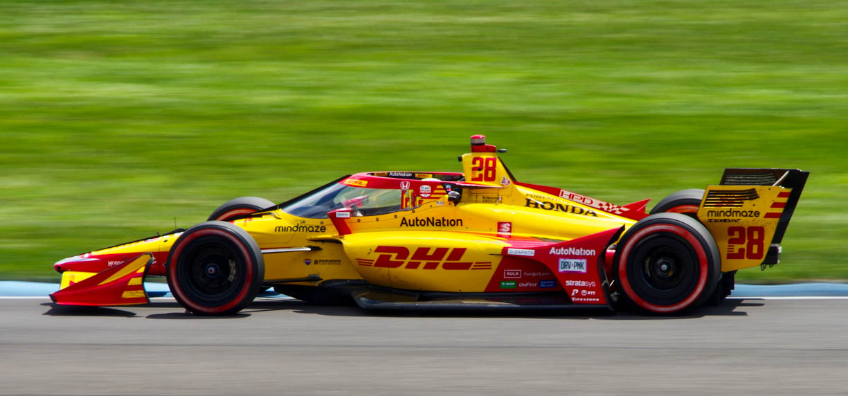 Romain Grosjean's 2022 Indycar racing at speeds over 120+mph during the 2nd 2022 Indycar Grand Prix at the Indianapolis Motor Speedway in central Indiana, USA. The race car is yellow and red and is moving very quickly around a right hand corner on the race track.