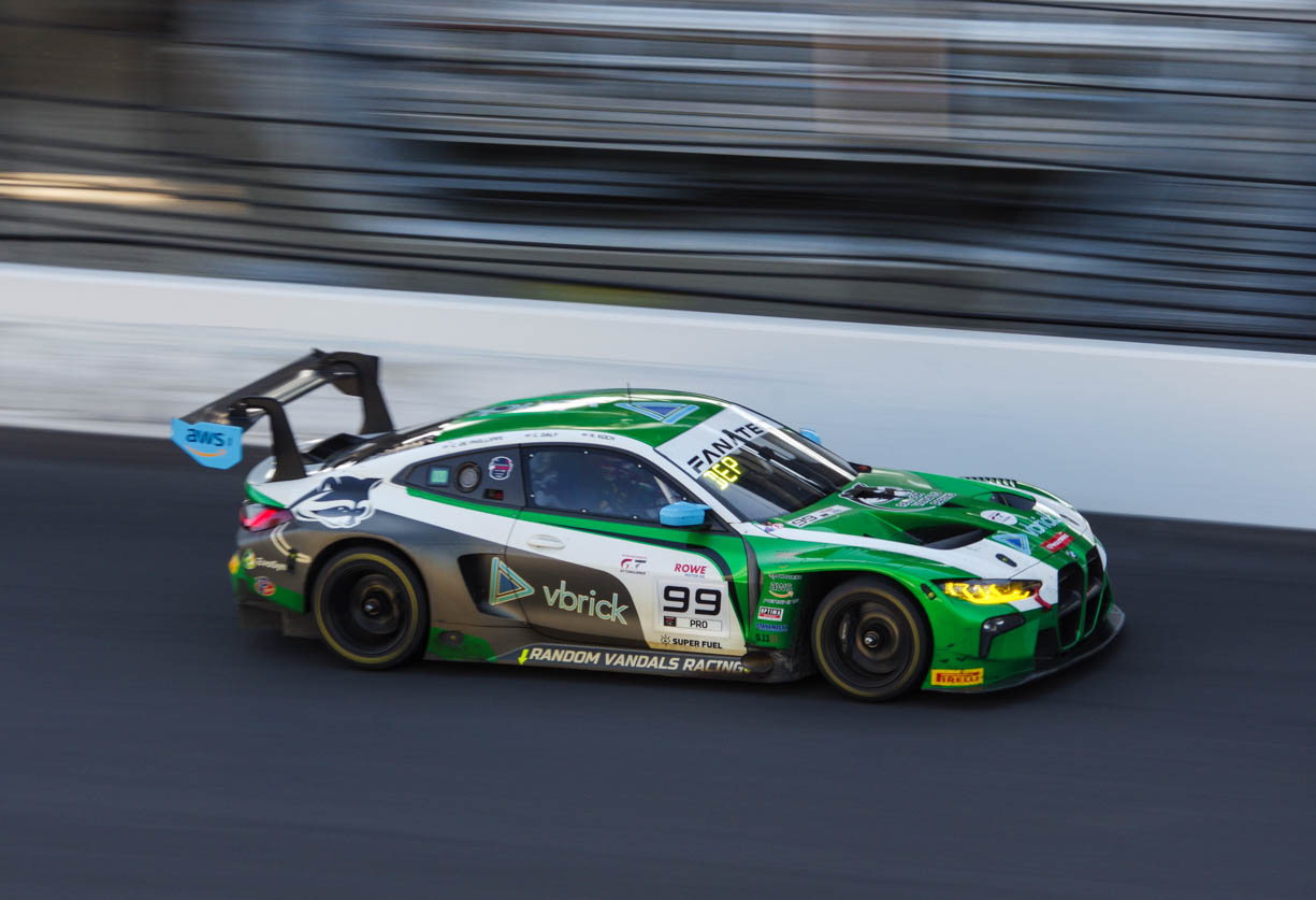 A green and white BMW M4 race car racing at high speeds down the front straight at the Indianapolis Motor Speedway during the 8 hour race. The image shows the car's headlights and number on the side of the car are illuminated which means that the photo is taken during dusk.