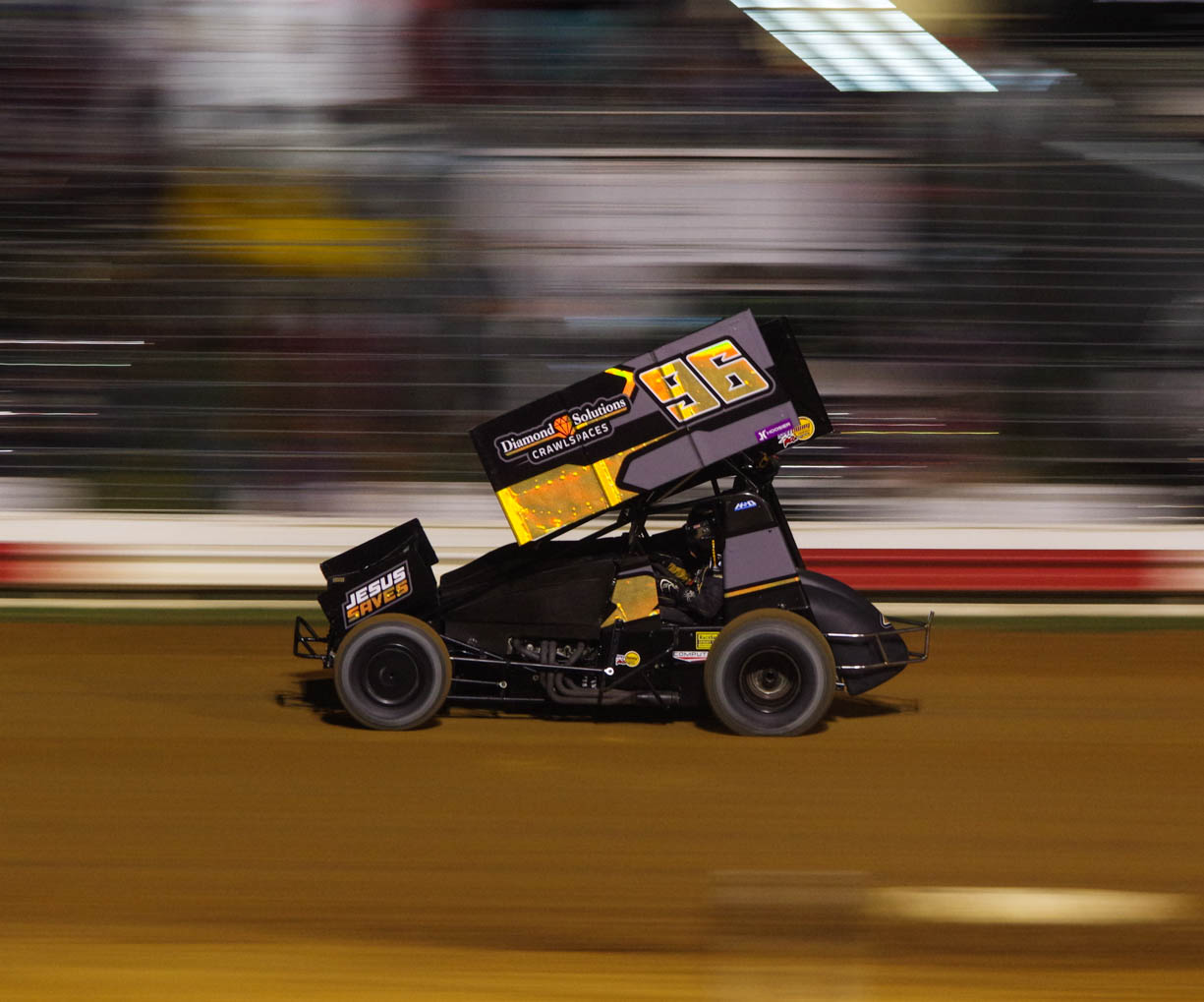 An orange and black 410 sprint car racing at high speeds along the back straight at the Paragon Speedway in central Indiana during the summer of 2024. The car is in focus, the background is blurry because the camera is panning to create a sense of speed. 