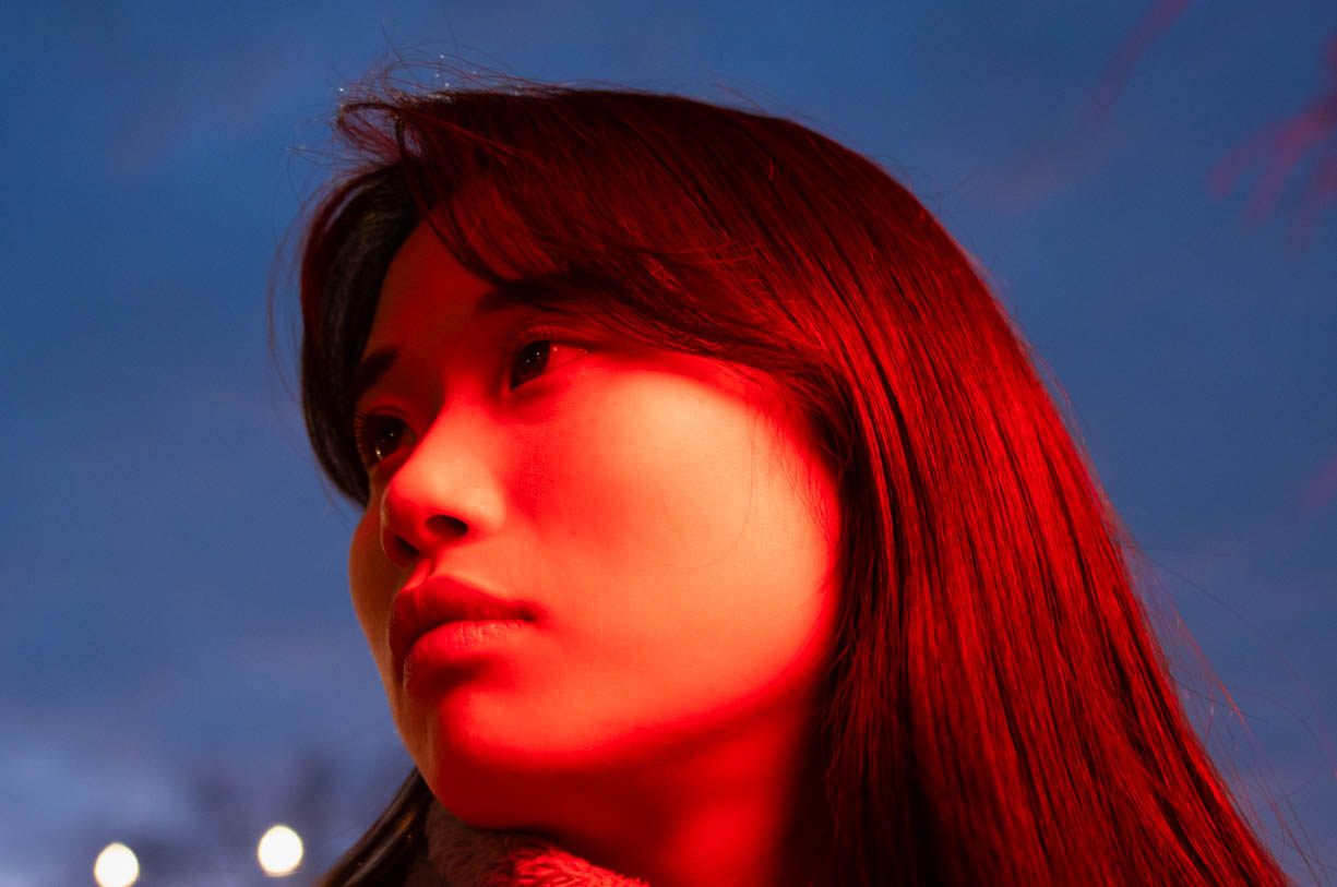 A woman photographed on the street in downtown Bloomington in 2021. The woman is illuminated by red neon signage at night to create the stark red color. 