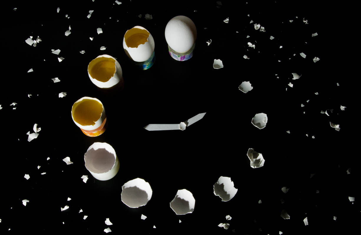 A clock made of broken eggs. Each hour of the clock is indicated by an egg, but the egg gets bigger as the hours progresses. At 9:00, the egg begins to have yolk in it and continues to get bigger, at 12:00 the egg is now a complete egg. The minute and hour hand are also made of eggs, plus there are shards of eggs scattered around the circular clock made of eggs. 