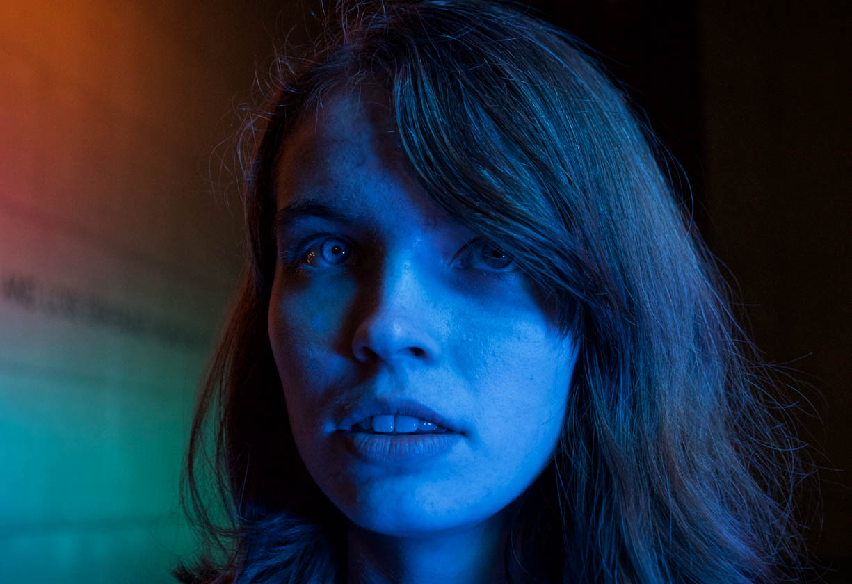 A photo of a beautiful woman that is illuminated by an intense colored outdoor light. The most intriguing aspect of this image are her eyes. They seem to be different colors (and different levels of brightness) because of the way the light source illuminates her face. The contrasts are so stark because there is a large LED colored light source that was used for this image. The LED light source was from a permanent structure like an outdoor art installation (the type that was used in this image). This image was taken during the fall of 2022. The woman is blue, one of her eyes is grey and one appears to be blue. Her hair is brown. There are red and green lights in the background on the left on the wall.