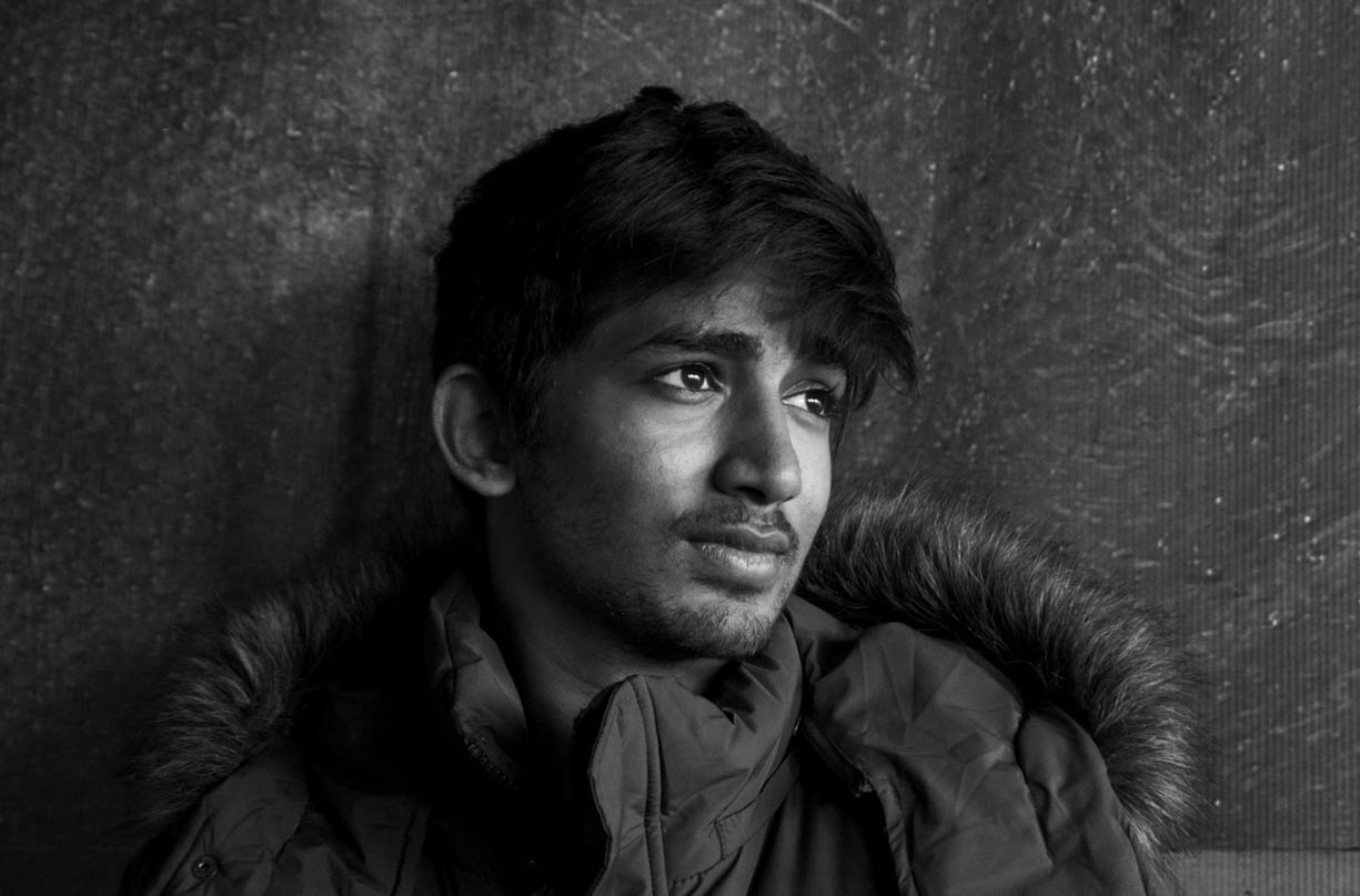 A young man photographed on the street in downtown Indianapolis near the convention center in black and white in 2021 for a university assignment. The young man has black hair and a winter jacket on that has fur around the hood. The image is also in black and white.