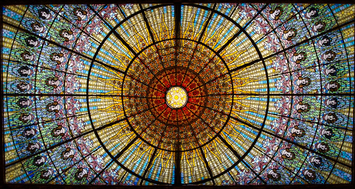 The spectacular stain glass window ceiling in the Palau de la Música Catalana in Barcelona, Spain. The stain glass window is red and orange in the middle like the sun, with blue, purple, and green surrounding it in a circular shape. Also around the sun-like shape are countless stain glass women with floral crowns. Both sides are symmetrical.