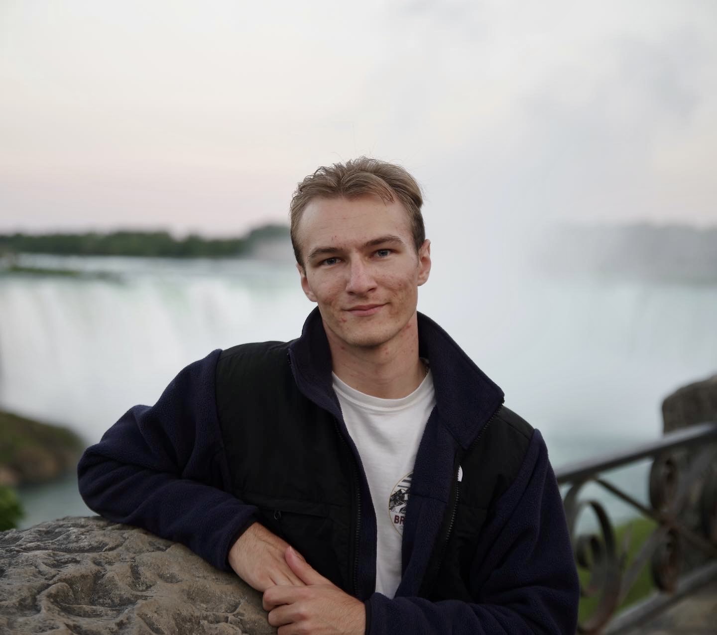 A photo of a good looking blonde haired man standing in front of Niagara Falls in Canada The man is looking at the camera.